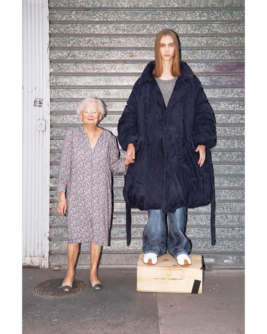 Two women, one elderly in a floral dress and the other younger in an oversized coat and jeans, stand outside a metal roll-up door.