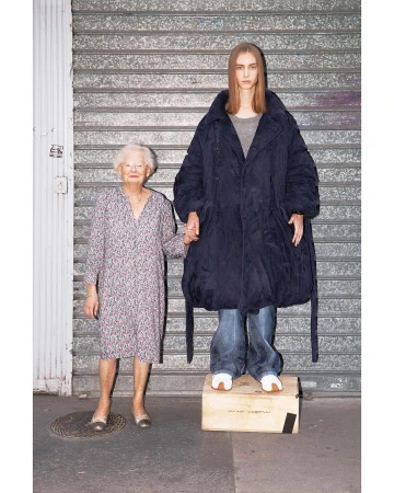 Two women, one elderly in a floral dress and the other younger in an oversized coat and jeans, stand outside a metal roll-up door.
