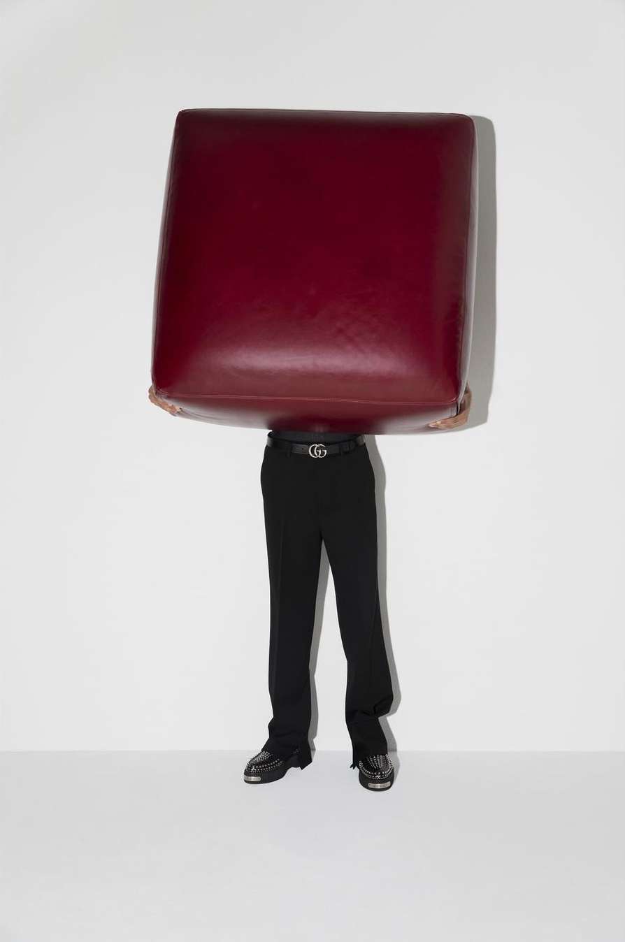 Person in black pants and shoes stands against a white wall, head hidden by a large burgundy, cube-shaped object held at head level.