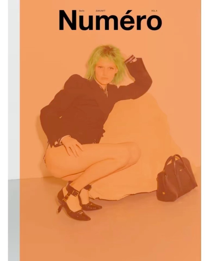 Magazine cover featuring a model with green hair, dark jacket, and heels, posing in a crouched position against an orange background.