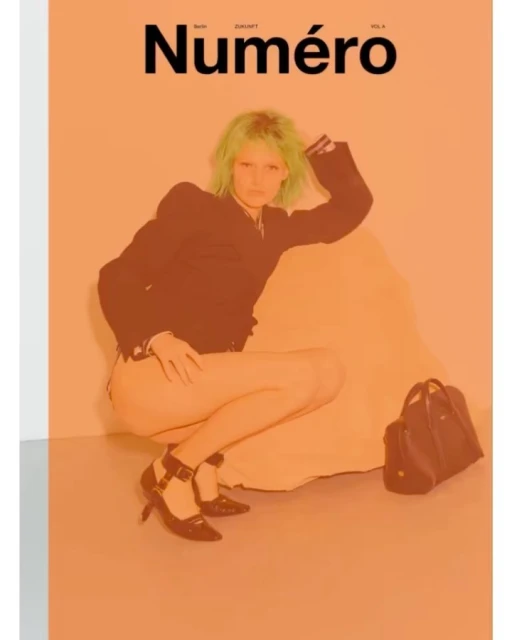 Magazine cover featuring a model with green hair, dark jacket, and heels, posing in a crouched position against an orange background.