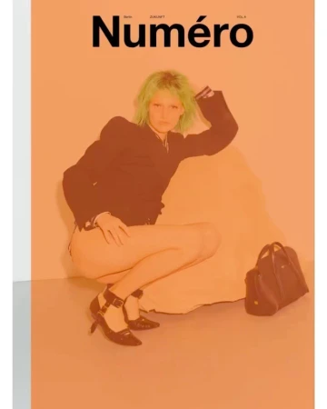 Magazine cover featuring a model with green hair, dark jacket, and heels, posing in a crouched position against an orange background.