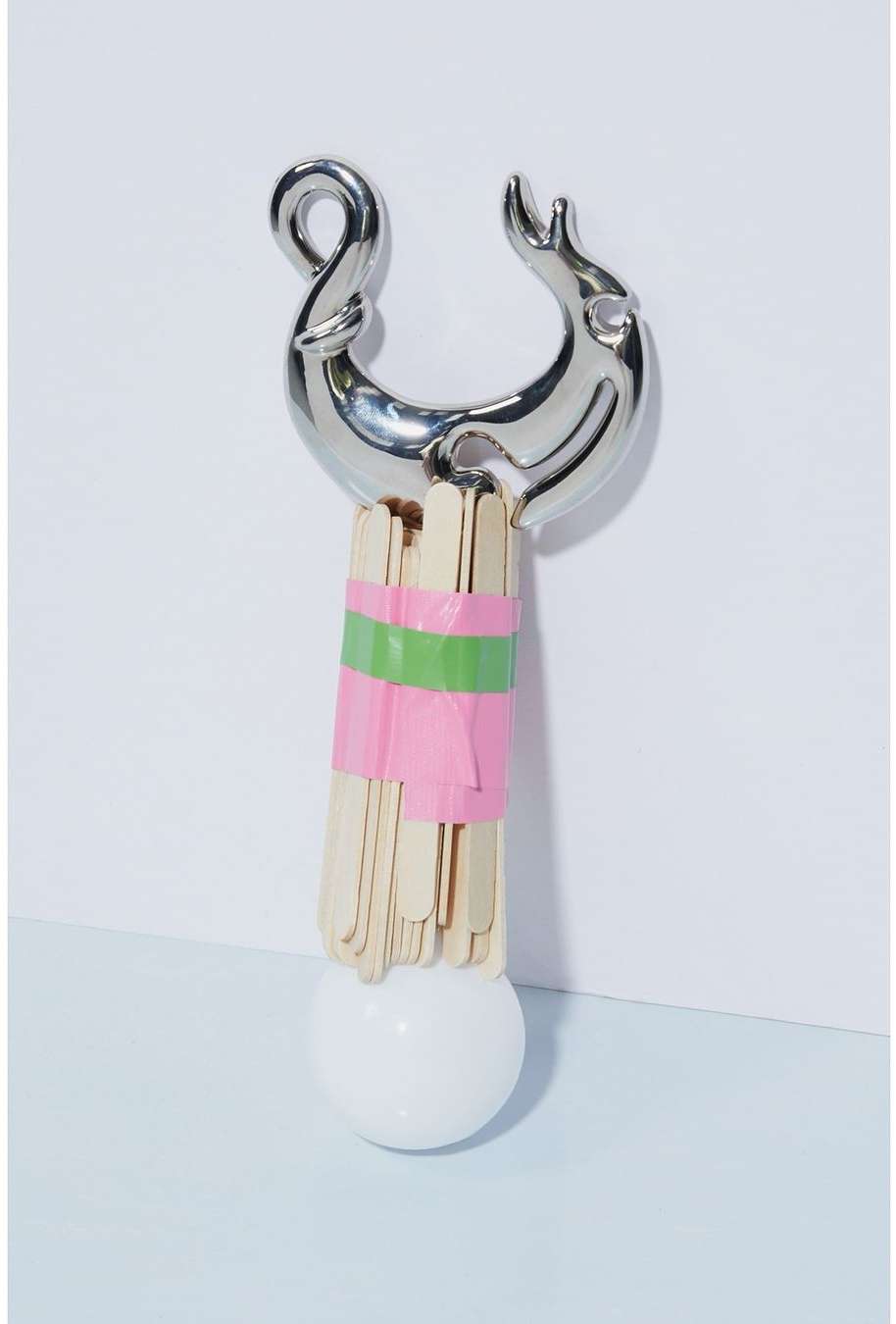 Abstract art piece featuring a chrome object, craft sticks wrapped with pink and green tape, and a white sphere against a white backdrop.