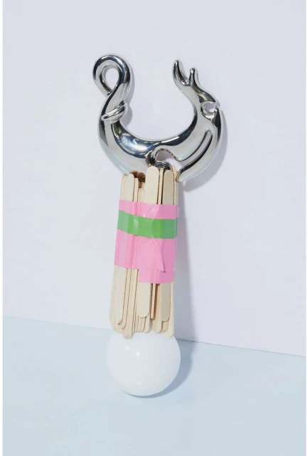 Abstract art piece featuring a chrome object, craft sticks wrapped with pink and green tape, and a white sphere against a white backdrop.