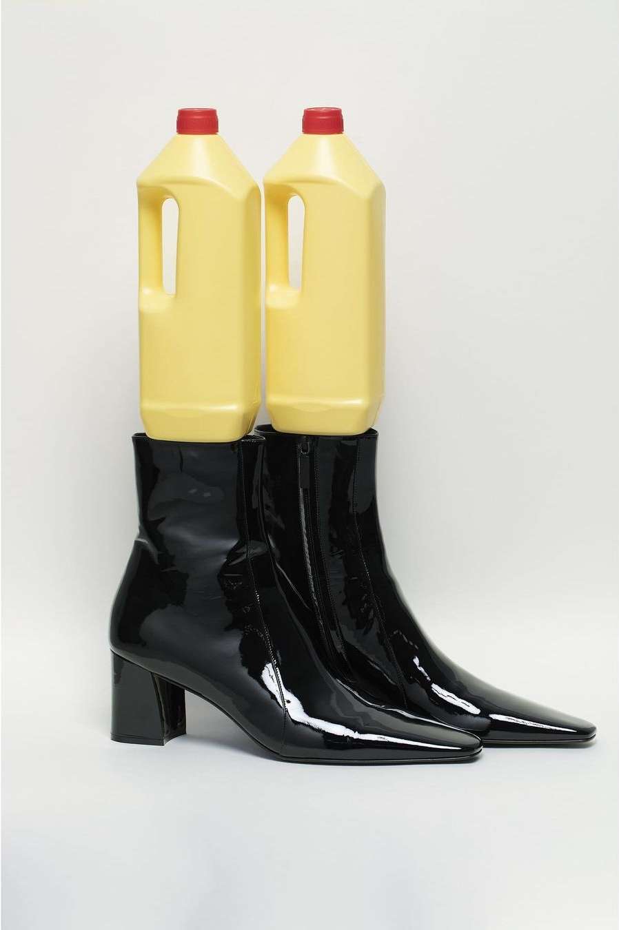 Two yellow cleaning bottles balance atop shiny black ankle boots in a studio shot on a white background, creating a surreal fashion image.