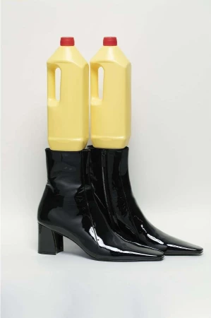 Two yellow cleaning bottles balance atop shiny black ankle boots in a studio shot on a white background, creating a surreal fashion image.