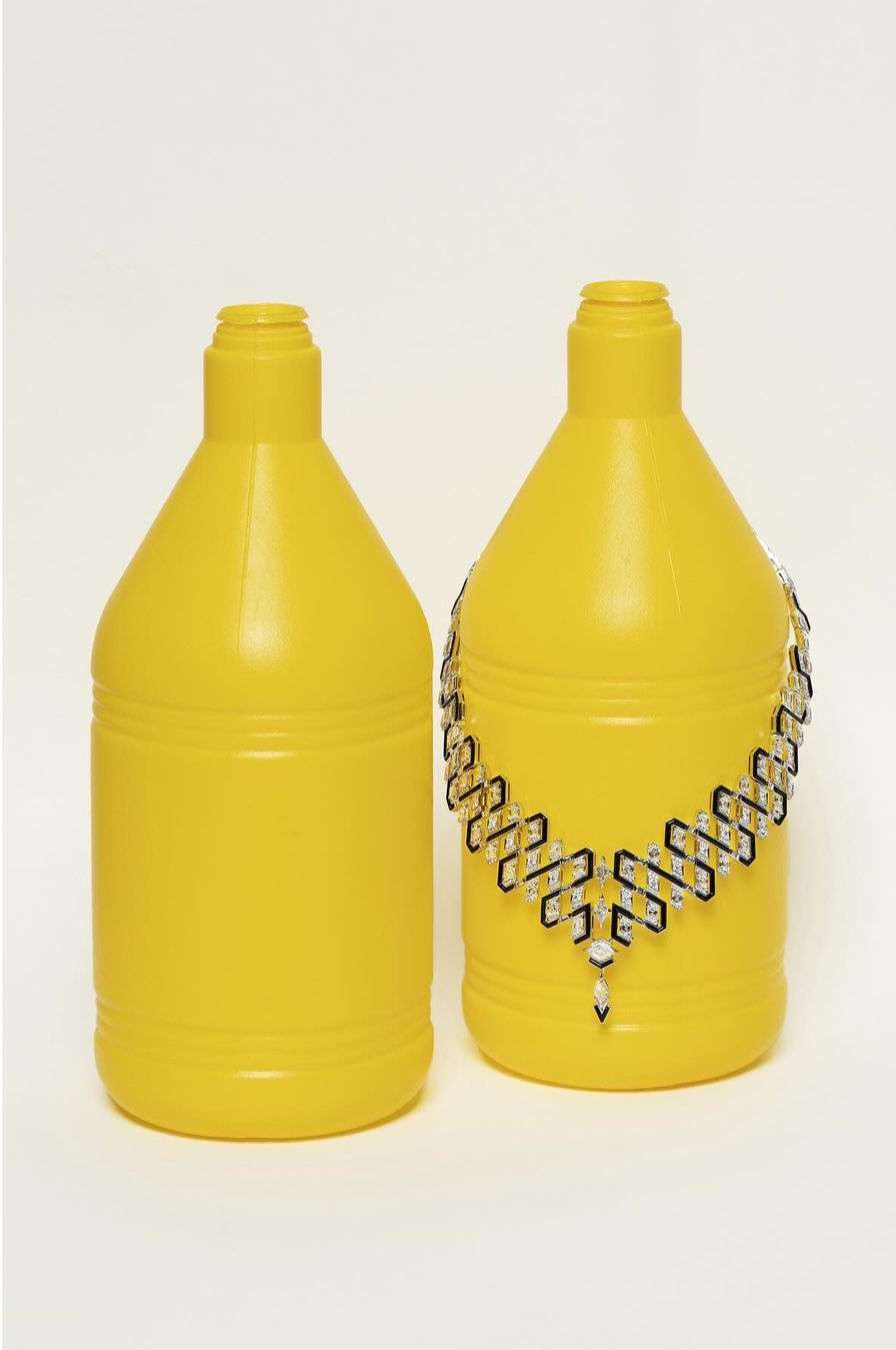 Two yellow plastic bottles are displayed indoors against a white background, one adorned with a decorative necklace.