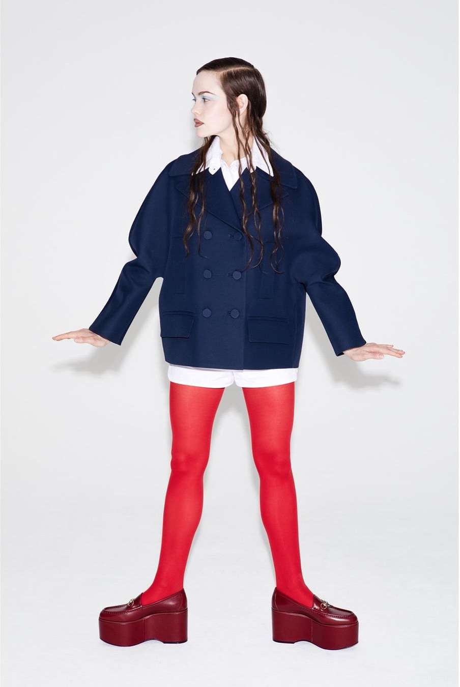 Fashion shot of a woman wearing a navy double-breasted blazer with red tights and platform loafers against a white backdrop.