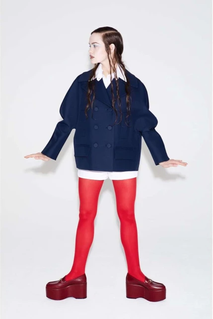 Fashion shot of a woman wearing a navy double-breasted blazer with red tights and platform loafers against a white backdrop.