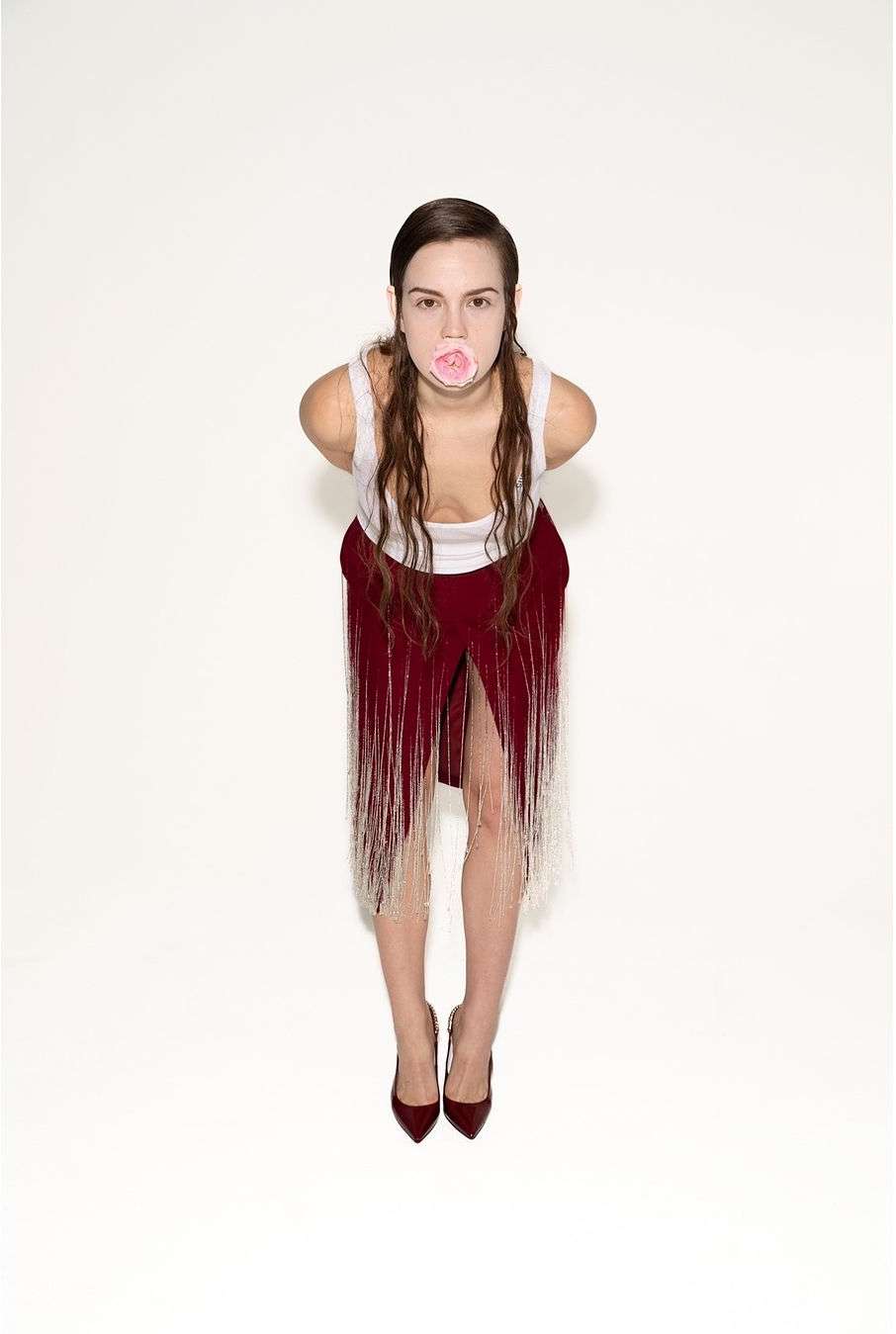 Full-length shot of a woman blowing a pink bubble, wearing a white top and a red fringed skirt against a white background. 