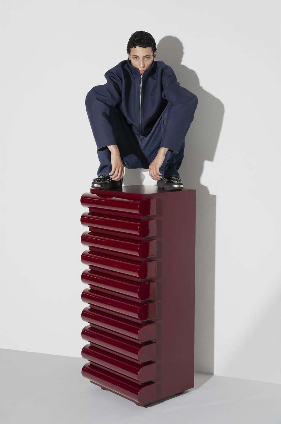 Fashionable adult male in a blue jumpsuit poses on a red, ridged cabinet in a studio setting with white walls and simple lighting.