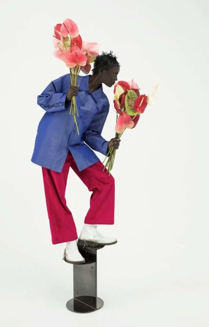 Fashion shot featuring an adult woman in a blue jacket and red pants stepping up onto a black pedestal while holding Anthurium flowers.
