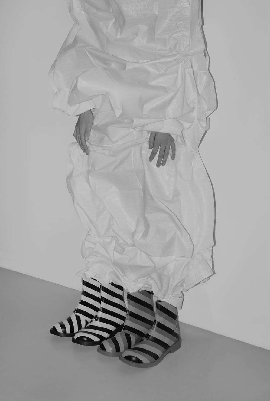 Person in white, oversized garment with striped boots against a neutral wall and floor, showcasing fashion and style.