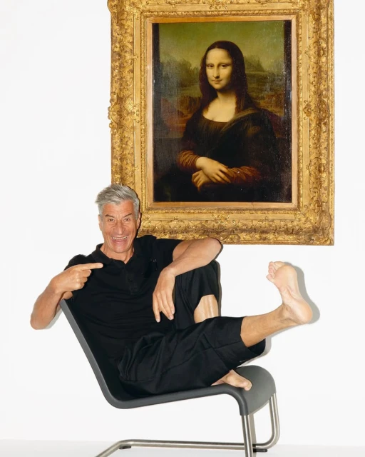 A man sits in a chair in front of a Mona Lisa painting in a white room. He is smiling and pointing towards the camera.