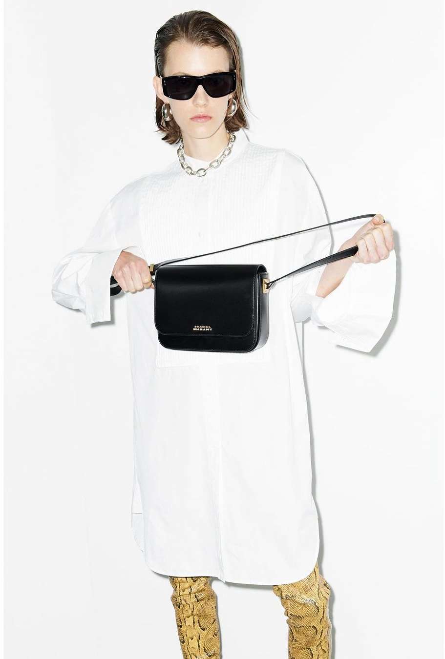 Woman with a black purse and sunglasses wearing a white oversized shirt and snake print boots in a fashion studio shot.
