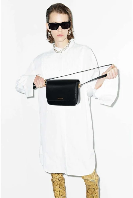 Woman with a black purse and sunglasses wearing a white oversized shirt and snake print boots in a fashion studio shot.