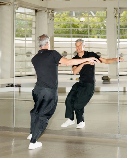 Man with grey hair in black shirt and pants in front of mirror in an open indoor space with light-colored walls and large windows.