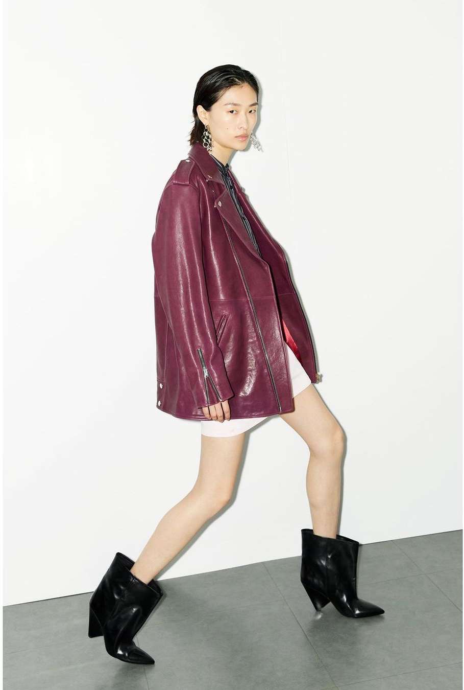 Fashion shot of a woman in a burgundy jacket and black boots walking in front of a white wall inside a photo studio.