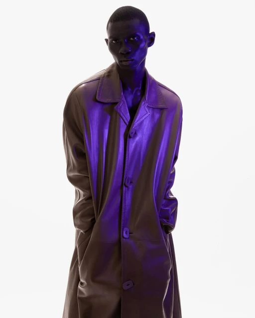 Fashion shot of a Black male model in a long brown coat, standing against a white background with blue studio lighting.