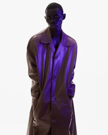 Fashion shot of a Black male model in a long brown coat, standing against a white background with blue studio lighting.