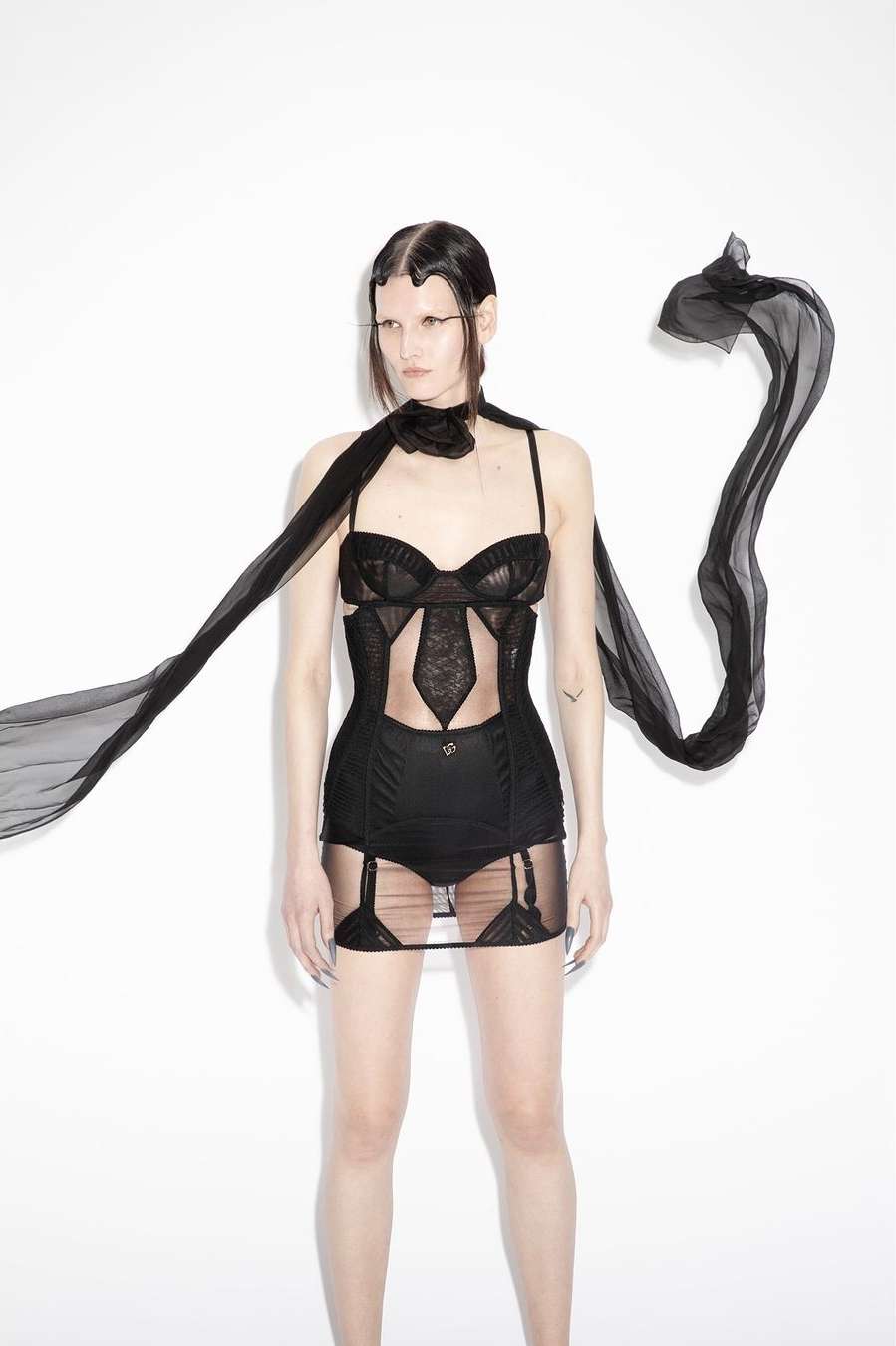 Adult woman poses in black lingerie with a sheer scarf on a white background in a studio. Fashion-forward clothing and long nails visible.