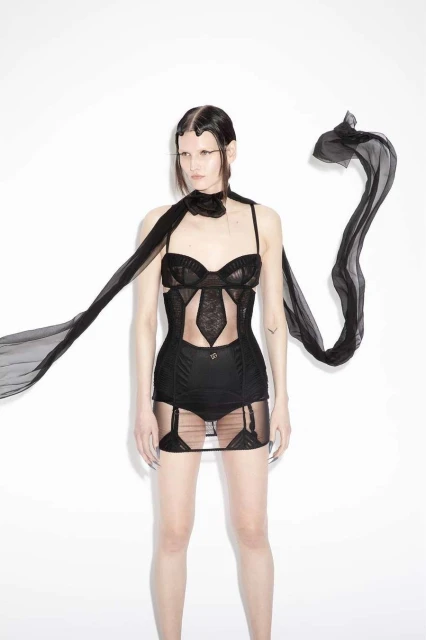 Adult woman poses in black lingerie with a sheer scarf on a white background in a studio. Fashion-forward clothing and long nails visible.