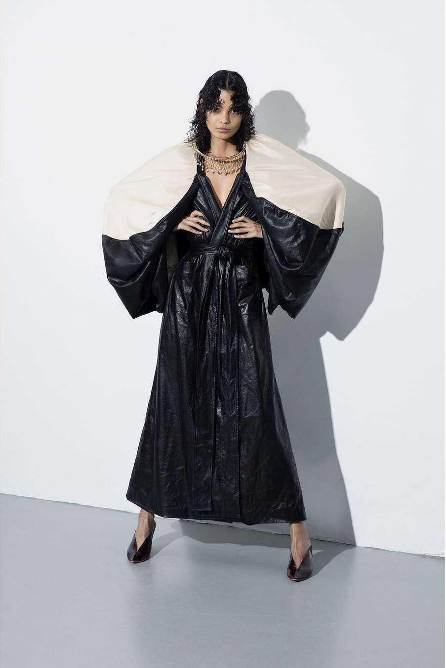 Woman in full-length black, glossy robe with ivory shoulder pieces poses in photo studio; she wears gold necklace and dark heels.