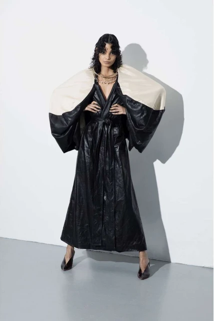 Woman in full-length black, glossy robe with ivory shoulder pieces poses in photo studio; she wears gold necklace and dark heels.