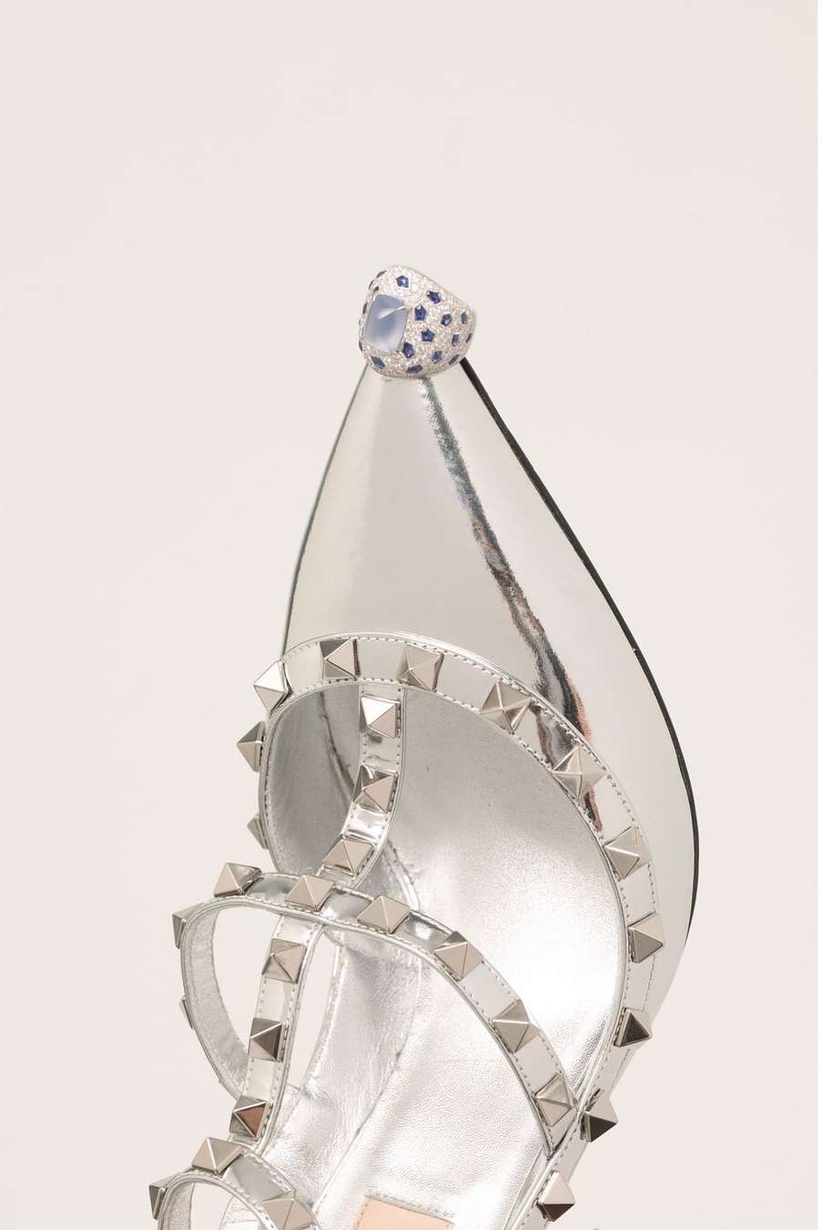 Close-up of a silver high-heel sandal with studded straps and a large jeweled ring on the toe, set against a white backdrop.