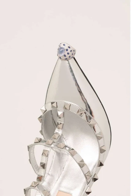 Close-up of a silver high-heel sandal with studded straps and a large jeweled ring on the toe, set against a white backdrop.