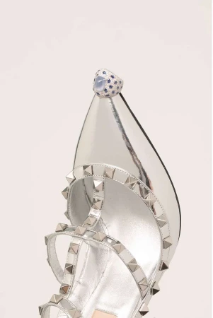 Close-up of a silver high-heel sandal with studded straps and a large jeweled ring on the toe, set against a white backdrop.
