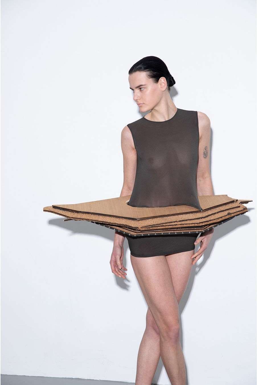 Woman poses in a sleeveless top and shorts with a cardboard skirt against a white backdrop in a studio fashion shoot.