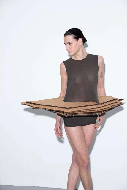 Woman poses in a sleeveless top and shorts with a cardboard skirt against a white backdrop in a studio fashion shoot.