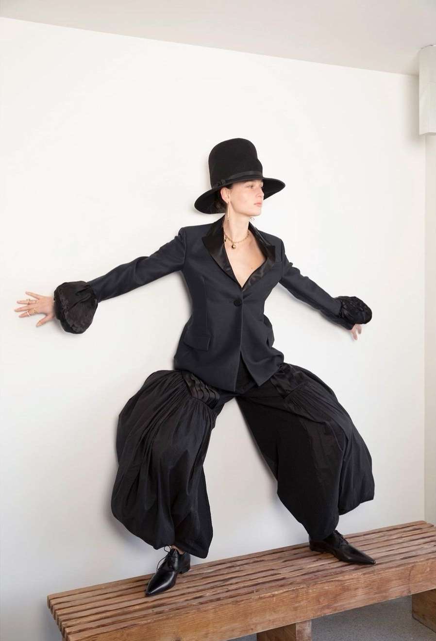 Fashionable woman in black top hat and oversized pants poses on bench against white wall, fashion-forward, avant-garde style.