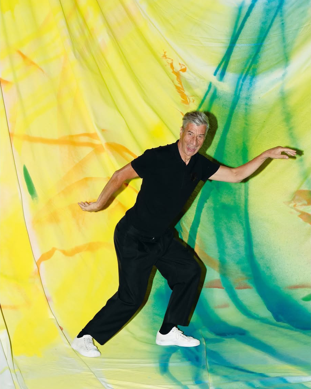 Older man in black outfit poses dynamically against a colorful, abstract backdrop, wearing white sneakers in a studio setting.