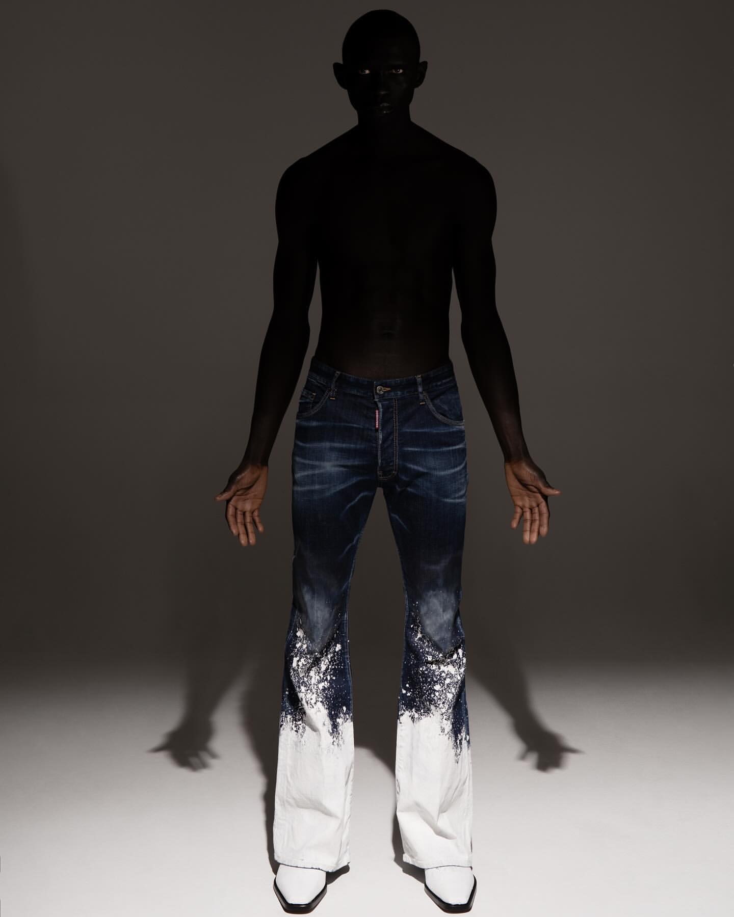 Fashion shot of a slender person with dark skin wearing dark blue denim pants that fade to white and pointy white shoes in a studio.