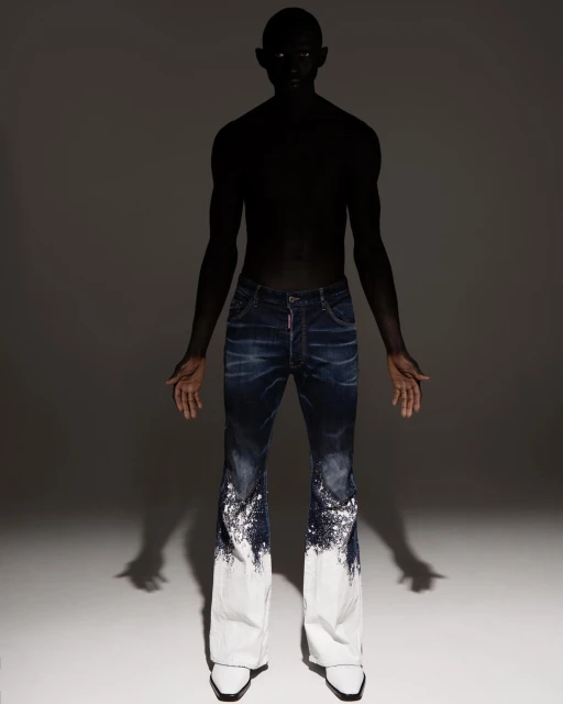 Fashion shot of a slender person with dark skin wearing dark blue denim pants that fade to white and pointy white shoes in a studio.