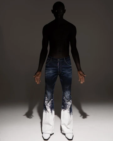 Fashion shot of a slender person with dark skin wearing dark blue denim pants that fade to white and pointy white shoes in a studio.