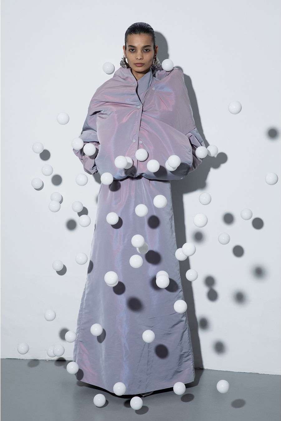A fashion shot features a woman in an iridescent lilac outfit standing in front of a white wall, surrounded by numerous white spheres.