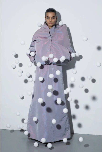 A fashion shot features a woman in an iridescent lilac outfit standing in front of a white wall, surrounded by numerous white spheres.