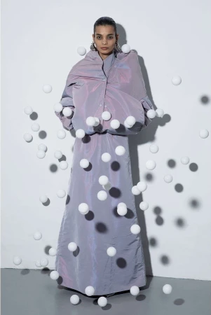 A fashion shot features a woman in an iridescent lilac outfit standing in front of a white wall, surrounded by numerous white spheres.