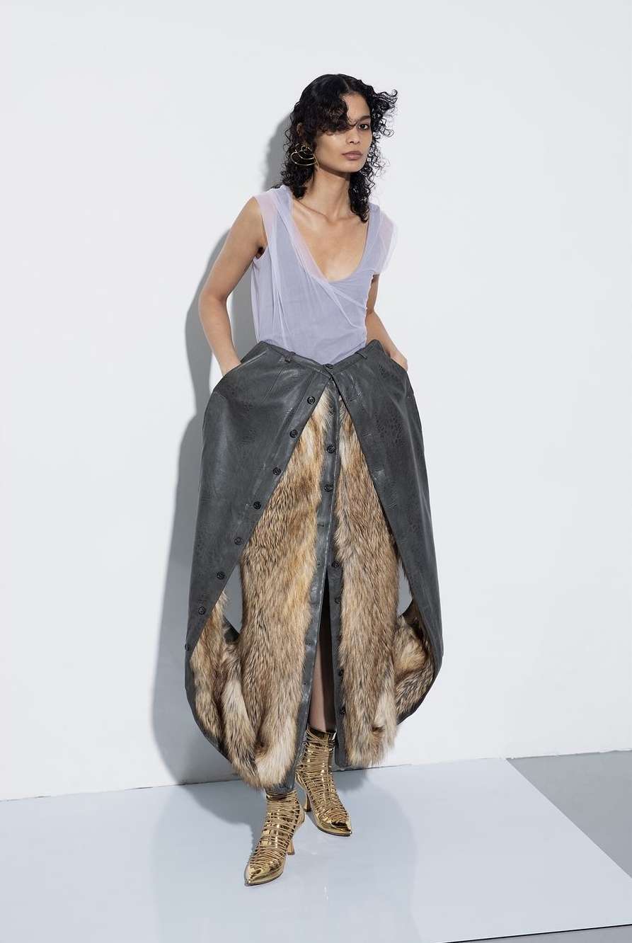 Fashion photo: A woman stands in a studio wearing a layered dress, featuring leather and fur, paired with metallic gold boots.