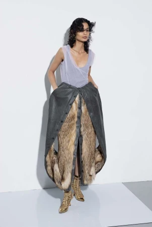 Fashion photo: A woman stands in a studio wearing a layered dress, featuring leather and fur, paired with metallic gold boots.