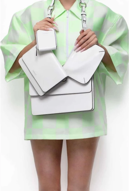 Woman showcasing a white leather shoulder bag with a unique flap design and a matching small coin purse, paired with a green-and-white dress.