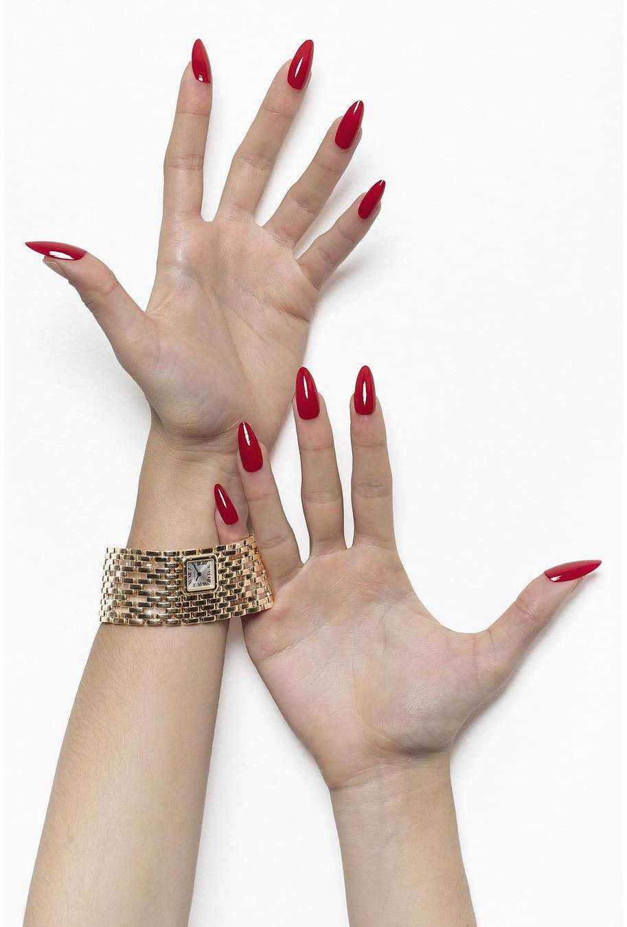 Two hands showcase long, almond-shaped nails painted in a bold red colour, accessorized with a gold watch against a white background.