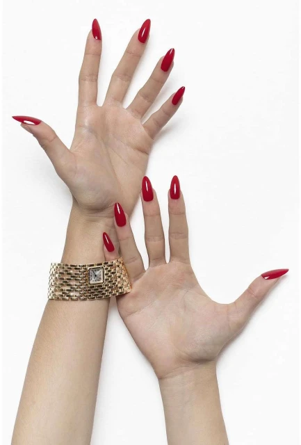 Two hands showcase long, almond-shaped nails painted in a bold red colour, accessorized with a gold watch against a white background.