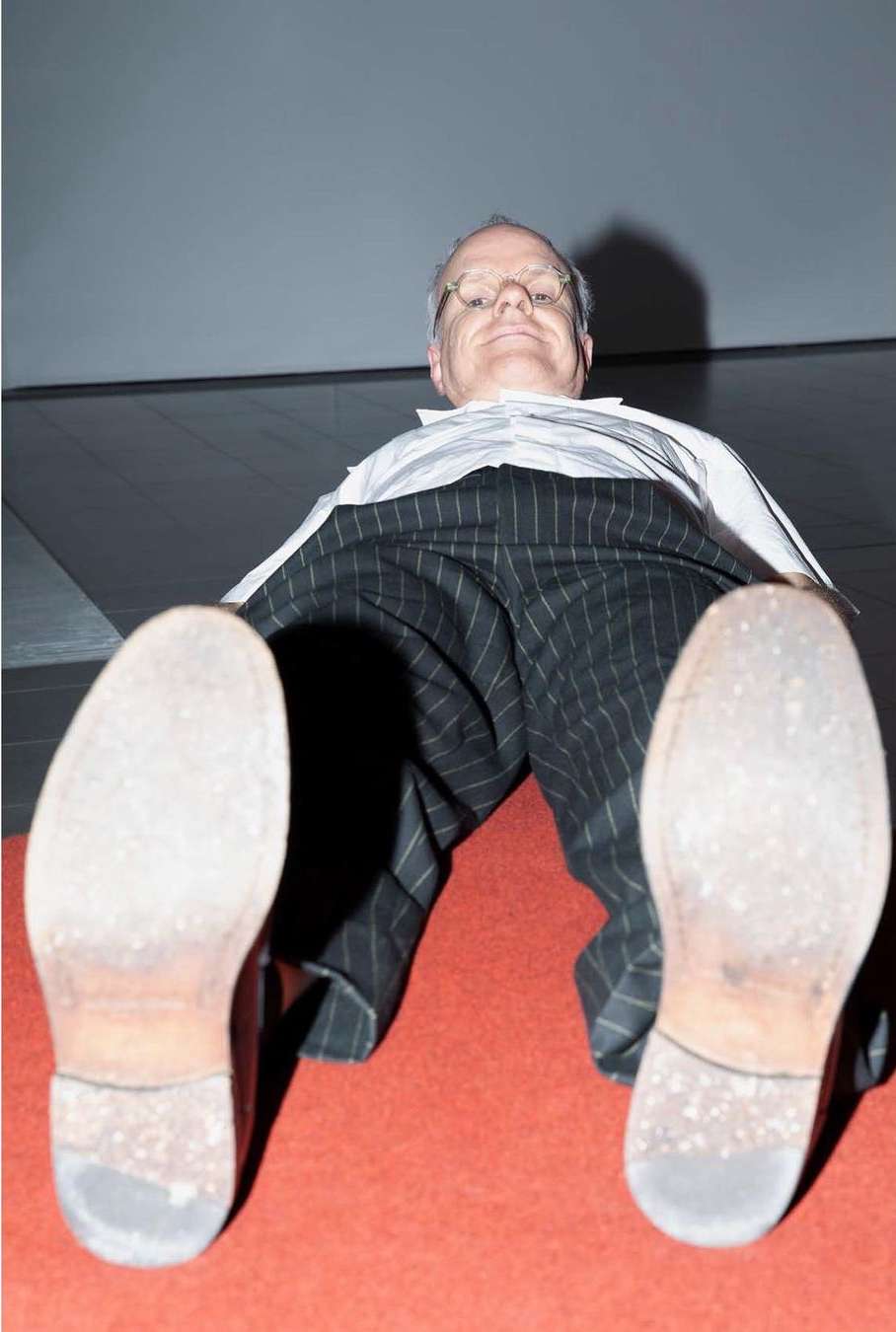 Low-angle shot of an adult male wearing pinstripe pants and dress shoes, lying on a red carpet under a grey ceiling, looking at the camera.