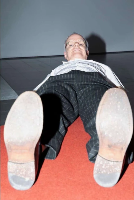 Low-angle shot of an adult male wearing pinstripe pants and dress shoes, lying on a red carpet under a grey ceiling, looking at the camera.