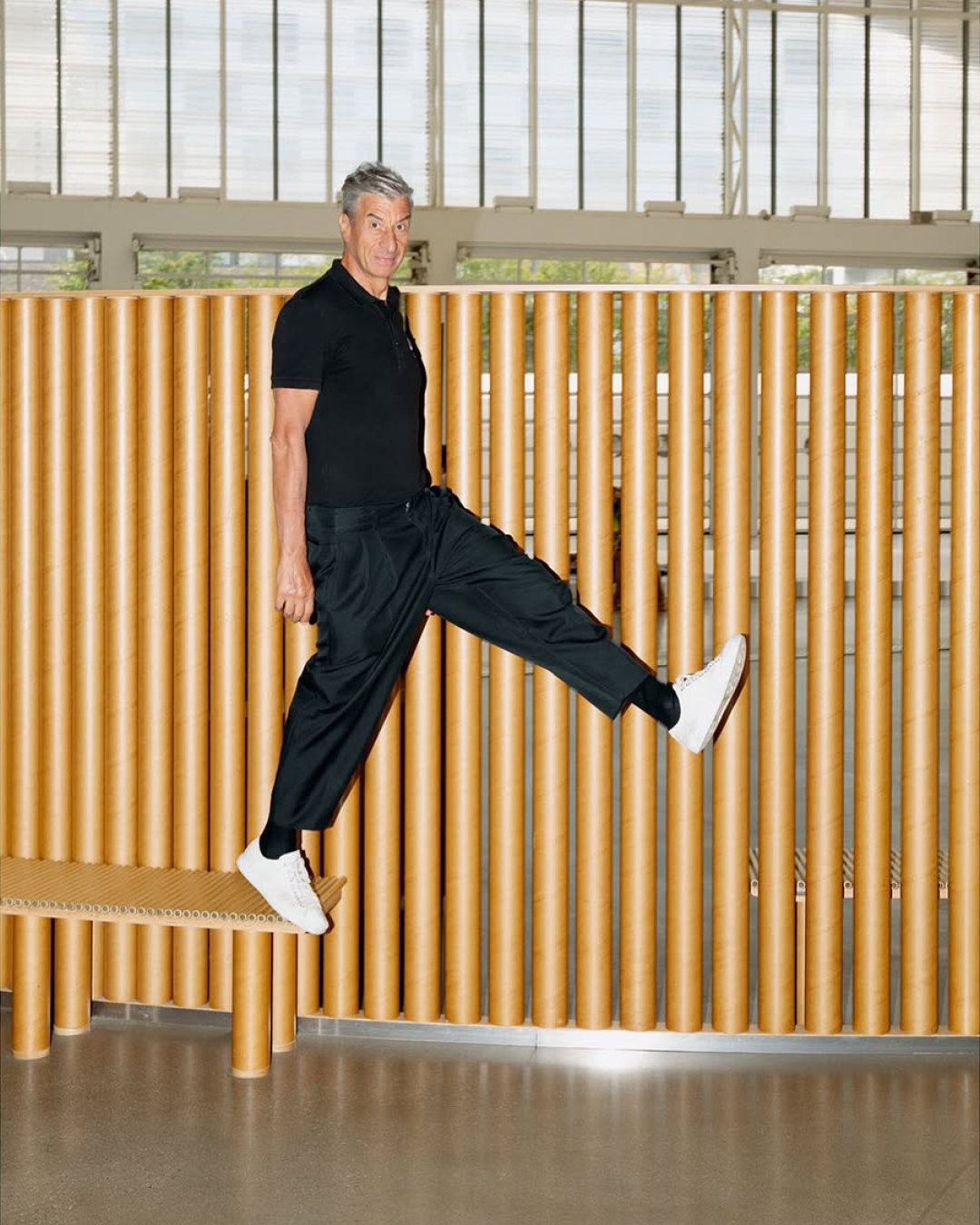 Adult man in a black polo shirt and black pants jumps off a bench, showcasing white sneakers in a brightly lit indoor setting.