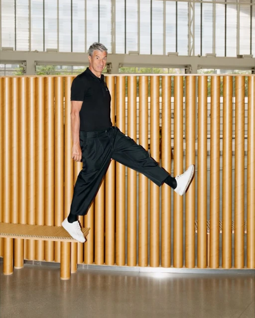 Adult man in a black polo shirt and black pants jumps off a bench, showcasing white sneakers in a brightly lit indoor setting.